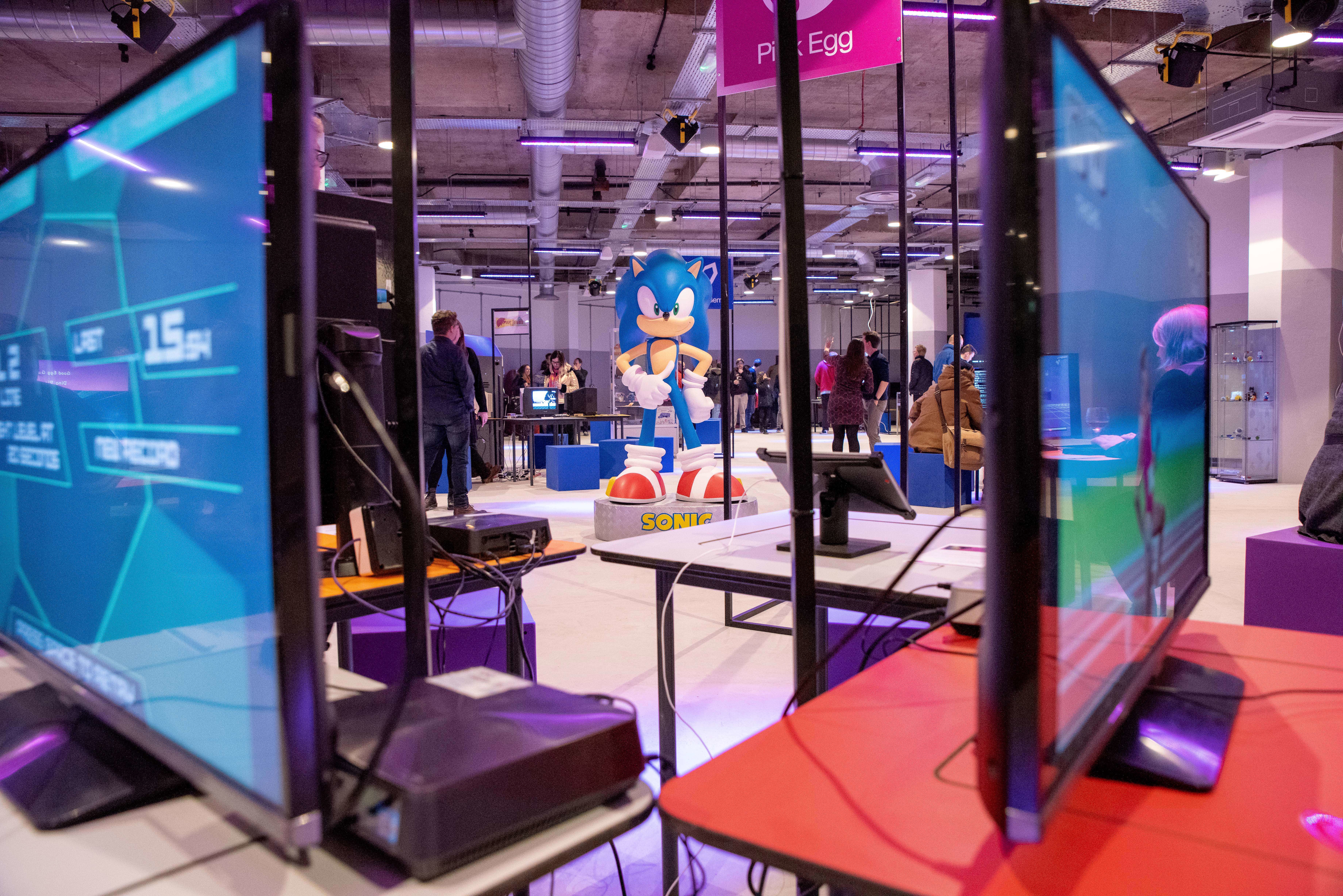 Interior of the National Videogame Museum with multiple gaming monitors in the foreground and a large Sonic the Hedgehog figure in the center. People are scattered around the space, interacting with exhibits. The room has an industrial ceiling with exposed pipes and bright lighting, creating a modern gaming environment.