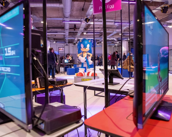 Interior of the National Videogame Museum with multiple gaming monitors in the foreground and a large Sonic the Hedgehog figure in the center. People are scattered around the space, interacting with exhibits. The room has an industrial ceiling with exposed pipes and bright lighting, creating a modern gaming environment.