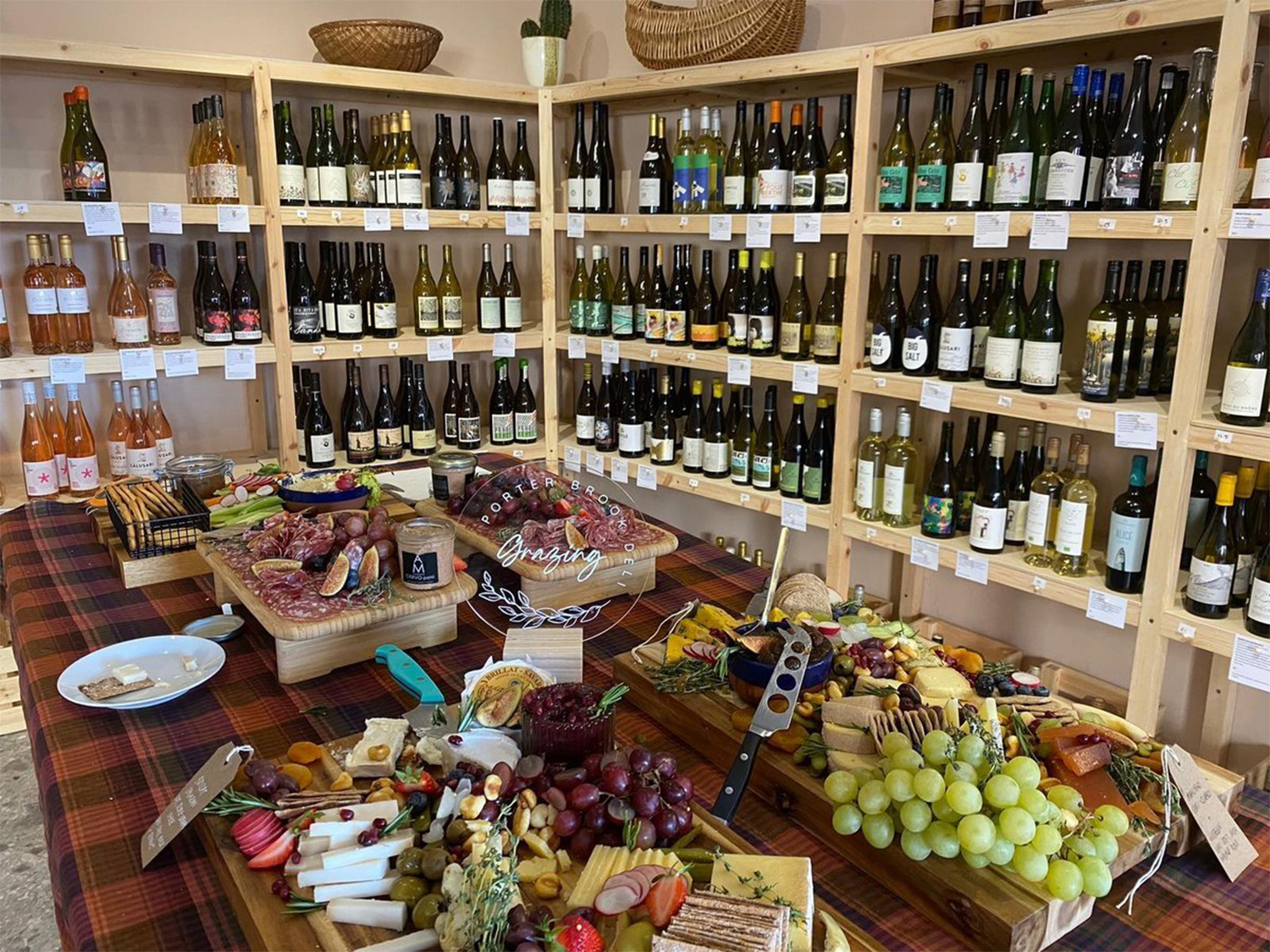 Rows of shelving filled with bottles of wine. In the centre of the room is a table covered in artfully arranged cheese, meats, grapes and other goods for sale.