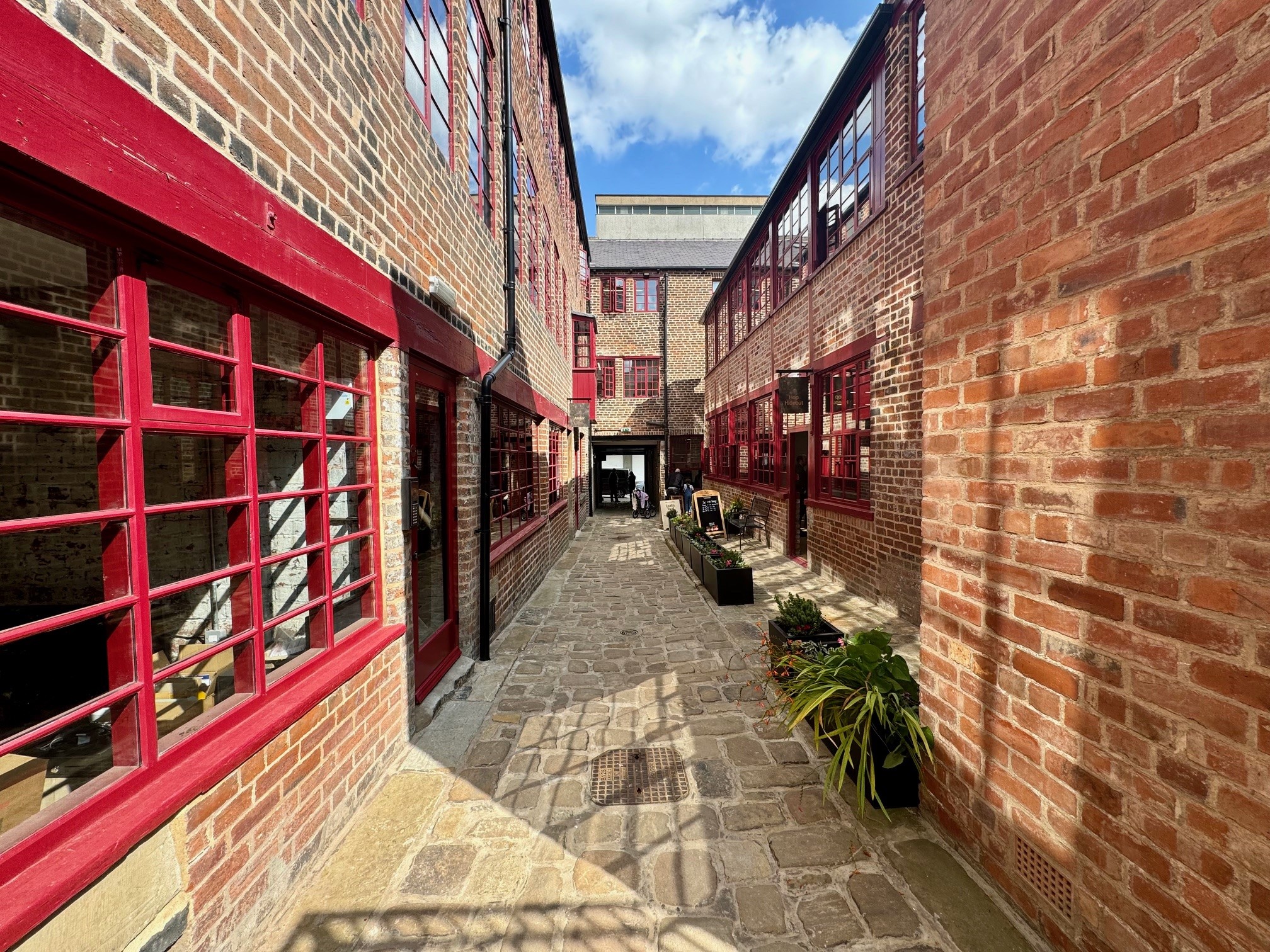 Narrow cobblestone alleyway between two red-brick industrial buildings with large red-framed windows. The passageway is lined with potted plants and benches, leading to an open doorway at the far end under a bright blue sky with scattered clouds.