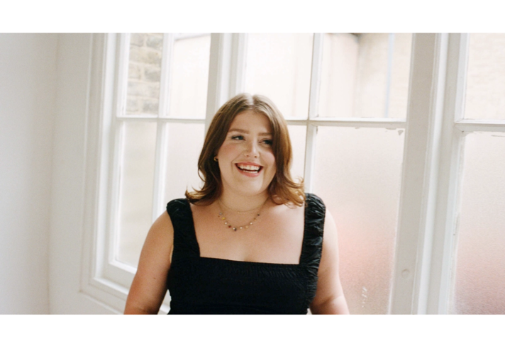 A woman, standing in front of a window, is laughing and looking happy.