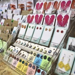 A close-up display of handmade jewellery including earrings, pins, and badges, arranged neatly on card backings with bright, colourful designs.