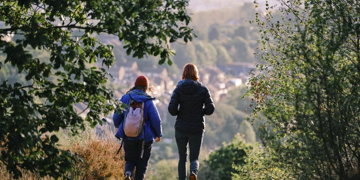 Two people walking along a dirt trail through a wooded area, wearing outdoor jackets and backpacks. The path leads downhill toward a scenic view of a town nestled among trees in the background. Sunlight creates a warm glow over the distant landscape, with green foliage framing the scene.