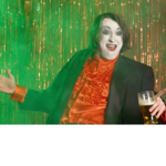 Person posing in front of a shimmering gold tinsel backdrop, lit with green stage-style lighting. Wearing a black jacket over a bright orange ruffled shirt, the person gestures with one hand while holding a pint glass in the other. The colourful, theatrical image has a cabaret or performance atmosphere, with metallic streamers filling the background.