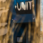 Exterior view of a blue-painted brick building with a black sign reading “UNT” above a recessed entrance. Tall golden grasses in the foreground partially obscure the view, creating a soft blurred frame. Sunlight casts shadows across the building, highlighting a large black-and-white mural on the wall, security lights, and architectural details. The roof of a parked car is visible at the bottom of the image.