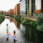 Three people are stand up paddle boarding on a canal that runs through a city centre.