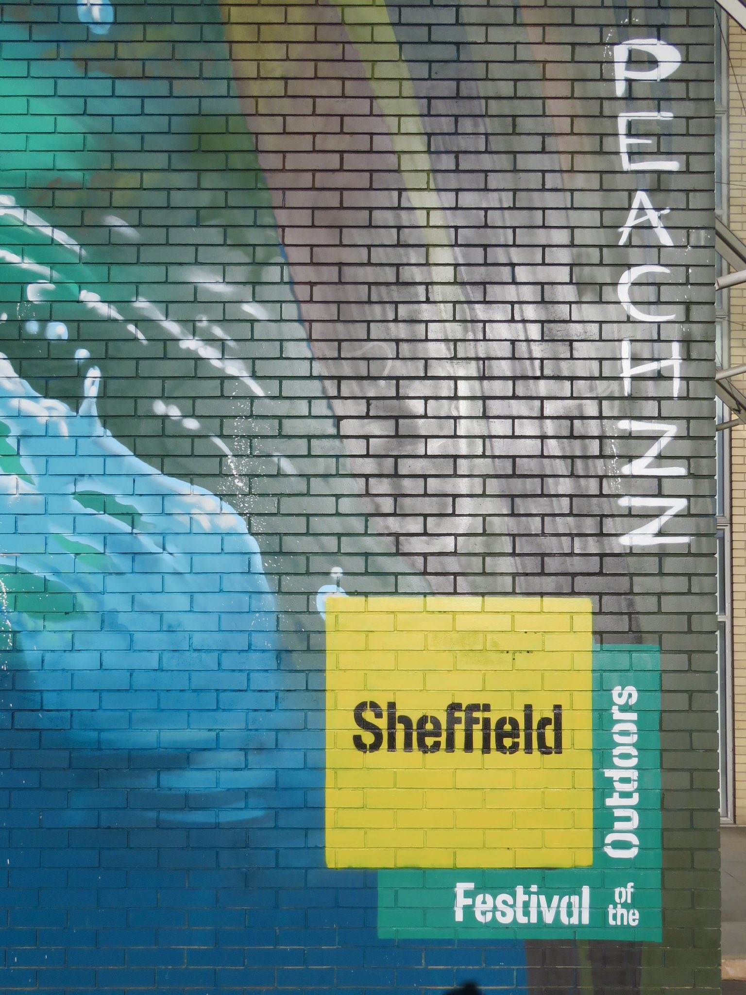 Close-up of a painted brick wall featuring part of a mural with blue and green water-like patterns. The word ‘PEACHZZ’ is written vertically in white letters on the right side. At the bottom, a yellow and green block of text reads ‘Sheffield Festival of the Outdoors .’