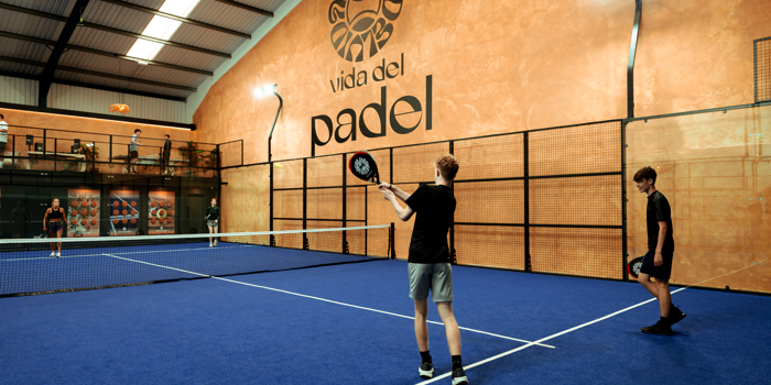 Indoor padel court with blue flooring, where two players prepare to rally while others watch from a raised viewing area against an orange wall displaying the ‘Vida del Padel’ logo