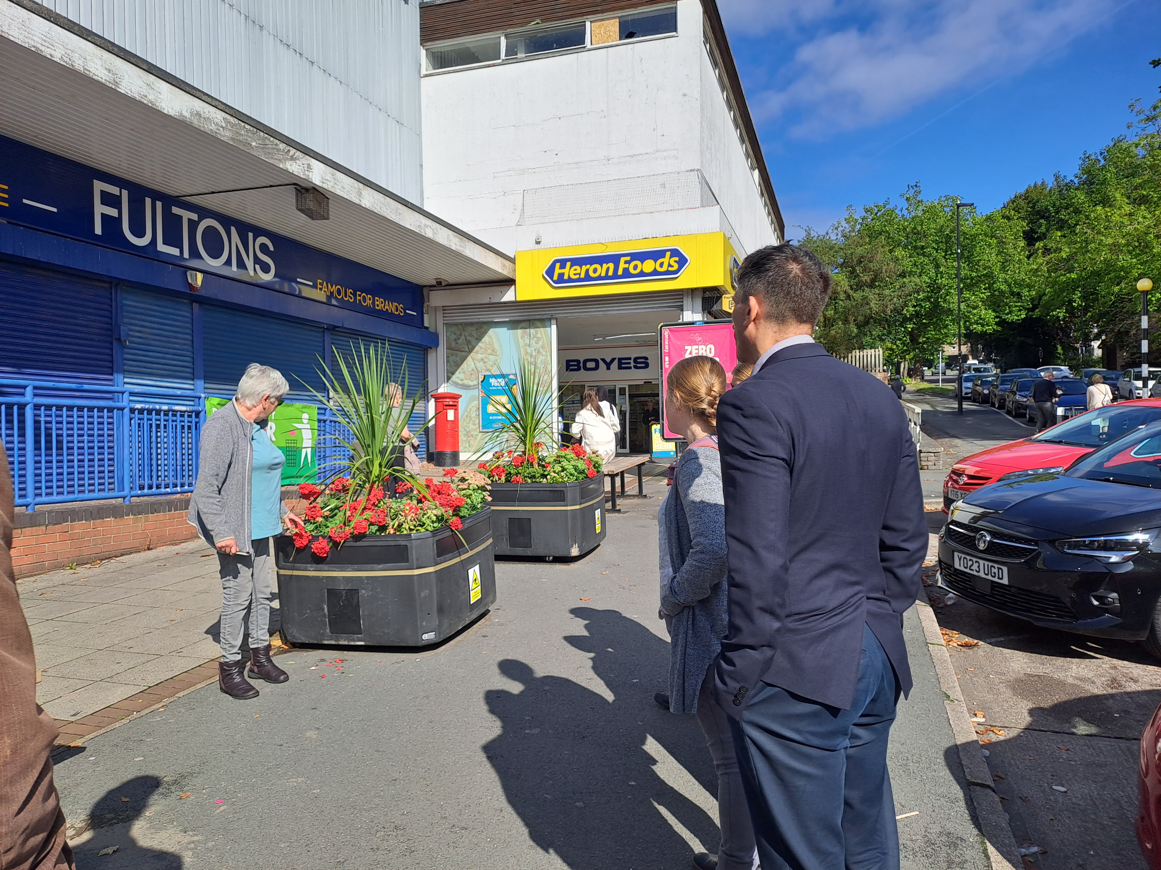 Street scene outside closed Fultons store and Heron Foods entrance, with large black planters filled with red flowers and green plants on the pavement. Several people are standing nearby, and parked cars line the road under a bright blue sky.