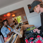 A person stands at a wooden record counter browsing vinyl records, while another operates DJ equipment with turntables and a mixer. Headphones and a colourful record sleeve are visible on the counter. The setting appears to be a cosy shop or café with white walls, hanging orange lights, shelves with bottles, and a tote bag displayed on the wall. A doorway in the background reveals more people and greenery outside.