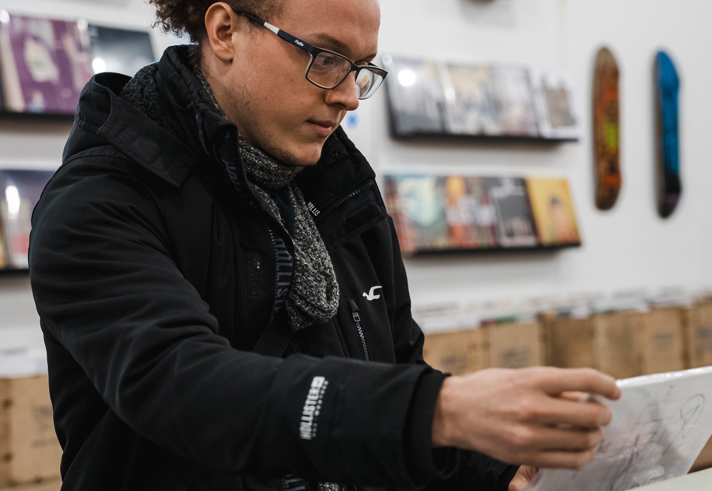 Person wearing a black jacket and patterned scarf, browsing through vinyl records in a record store. The foreground shows rows of records in bins with visible labels, and the background has shelves displaying album covers and a few skateboard decks mounted on the wall. The store has bright lighting and a modern interior.