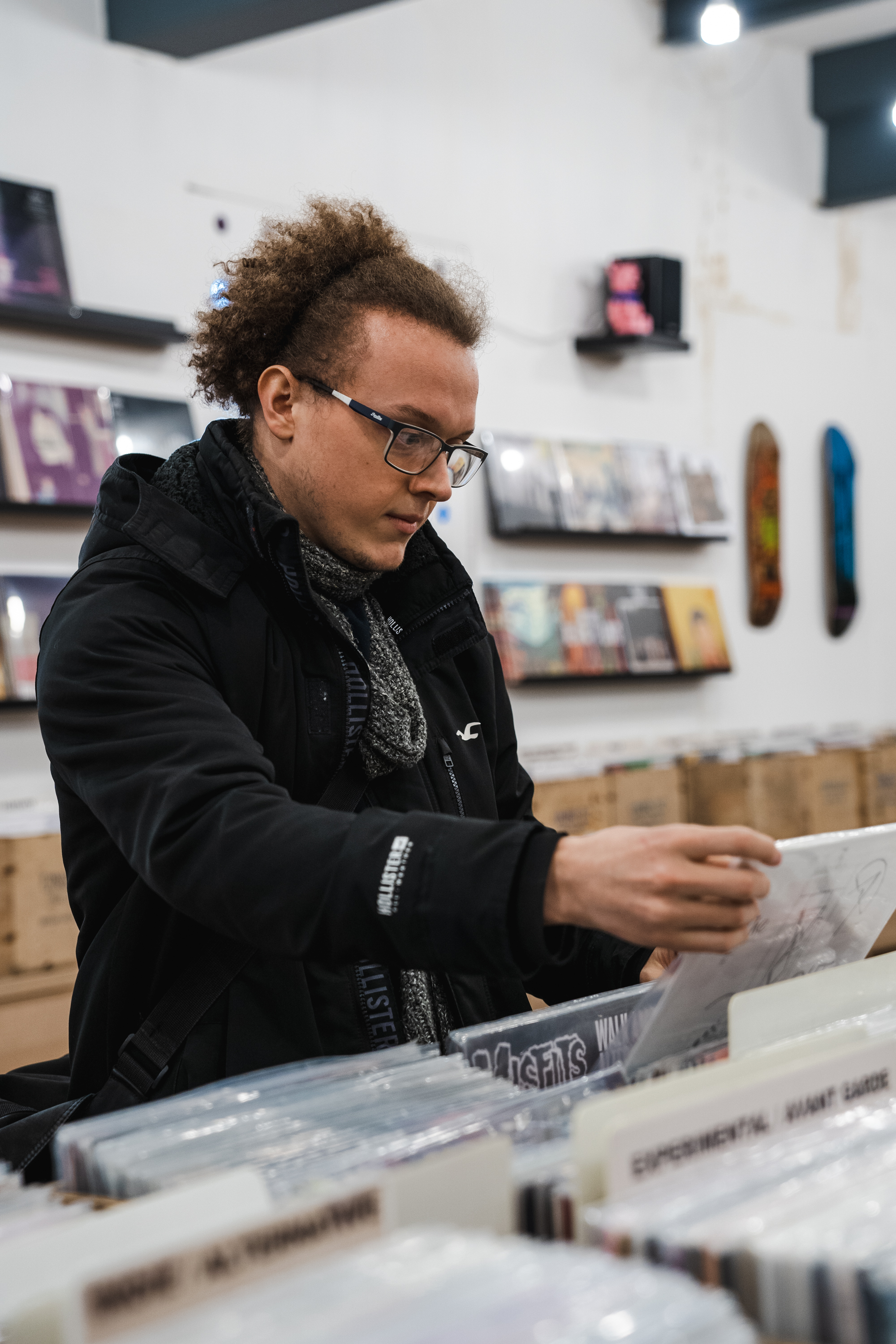 Person wearing a black jacket and patterned scarf, browsing through vinyl records in a record store. The foreground shows rows of records in bins with visible labels, and the background has shelves displaying album covers and a few skateboard decks mounted on the wall. The store has bright lighting and a modern interior.