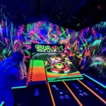 Woman taking a shot at on an indoor crazy golf course that looks like a giant pin ball machine and is illuminated with ultra violet lights 