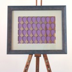 Framed artwork with a rustic wooden frame featuring a grid of purple teardrop shapes on a metallic background, displayed on an easel against a neutral wall.