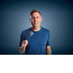 Promotional-style portrait of a person standing against a blue gradient background, holding a microphone in one hand and facing the camera. The person wears a plain blue T-shirt and is centred in the frame. Studio lighting highlights the figure, with a dark blue vignette around the edges creating a simple, professional backdrop.
