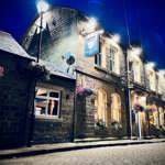 The Old Horns Inn at High Bradfield illuminated at night, with warm lights glowing from its stone-built exterior and hanging flower baskets decorating the front.