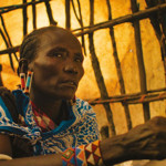 Close-up photograph of a person seated inside a structure made from wooden poles and woven material. The person wears a blue patterned shawl, colourful beaded earrings and matching beaded bracelets. Warm golden light fills the background, highlighting the texture of the shelter walls. The image focuses on traditional clothing and jewellery, with the person's hands resting in the foreground.