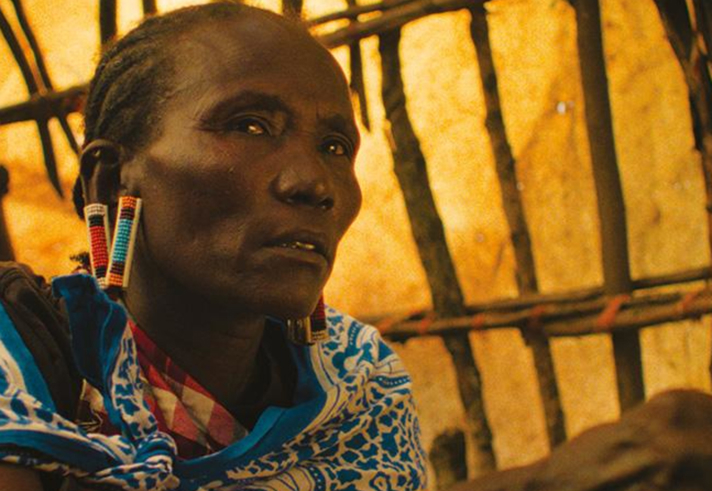 Close-up photograph of a person seated inside a structure made from wooden poles and woven material. The person wears a blue patterned shawl, colourful beaded earrings and matching beaded bracelets. Warm golden light fills the background, highlighting the texture of the shelter walls. The image focuses on traditional clothing and jewellery, with the person's hands resting in the foreground.