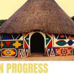 A traditional round building with a large thatched roof stands in a sandy landscape. Its walls are painted with bright geometric patterns in red, blue, yellow, green and black, creating a striking decorative design. A person sits on a low wall to the left of the building. The words “WORK IN PROGRESS” appear in large yellow text across the bottom of the image.