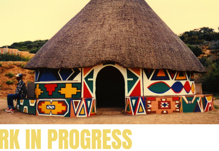 A traditional round building with a large thatched roof stands in a sandy landscape. Its walls are painted with bright geometric patterns in red, blue, yellow, green and black, creating a striking decorative design. A person sits on a low wall to the left of the building. The words “WORK IN PROGRESS” appear in large yellow text across the bottom of the image.