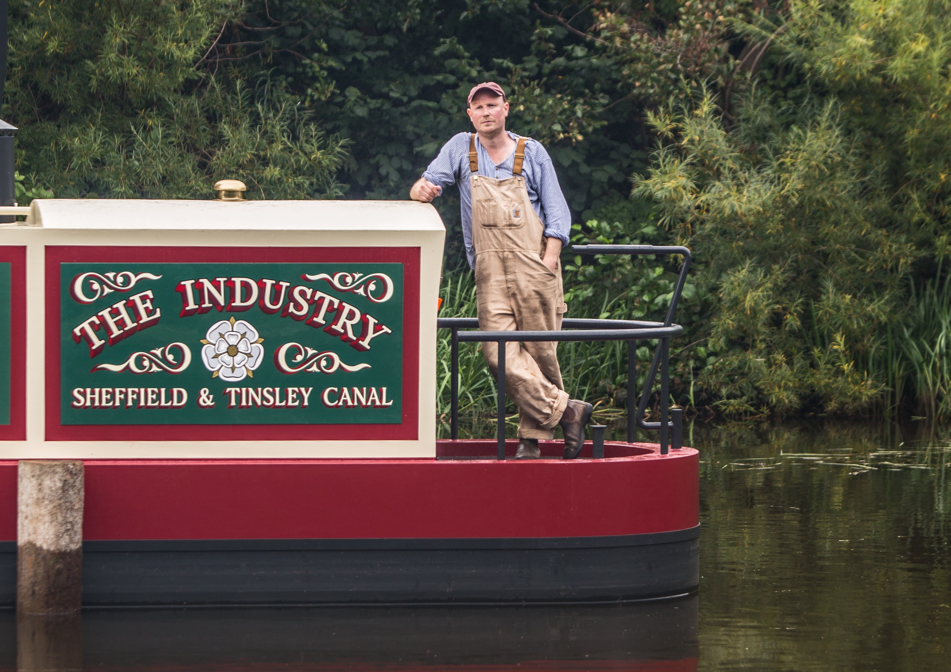 Person wearing light-colored overalls and a blue shirt leaning against a canal boat with a decorative sign reading ‘THE INDUSTRY Sheffield & Tinsley Canal,’ surrounded by green foliage.