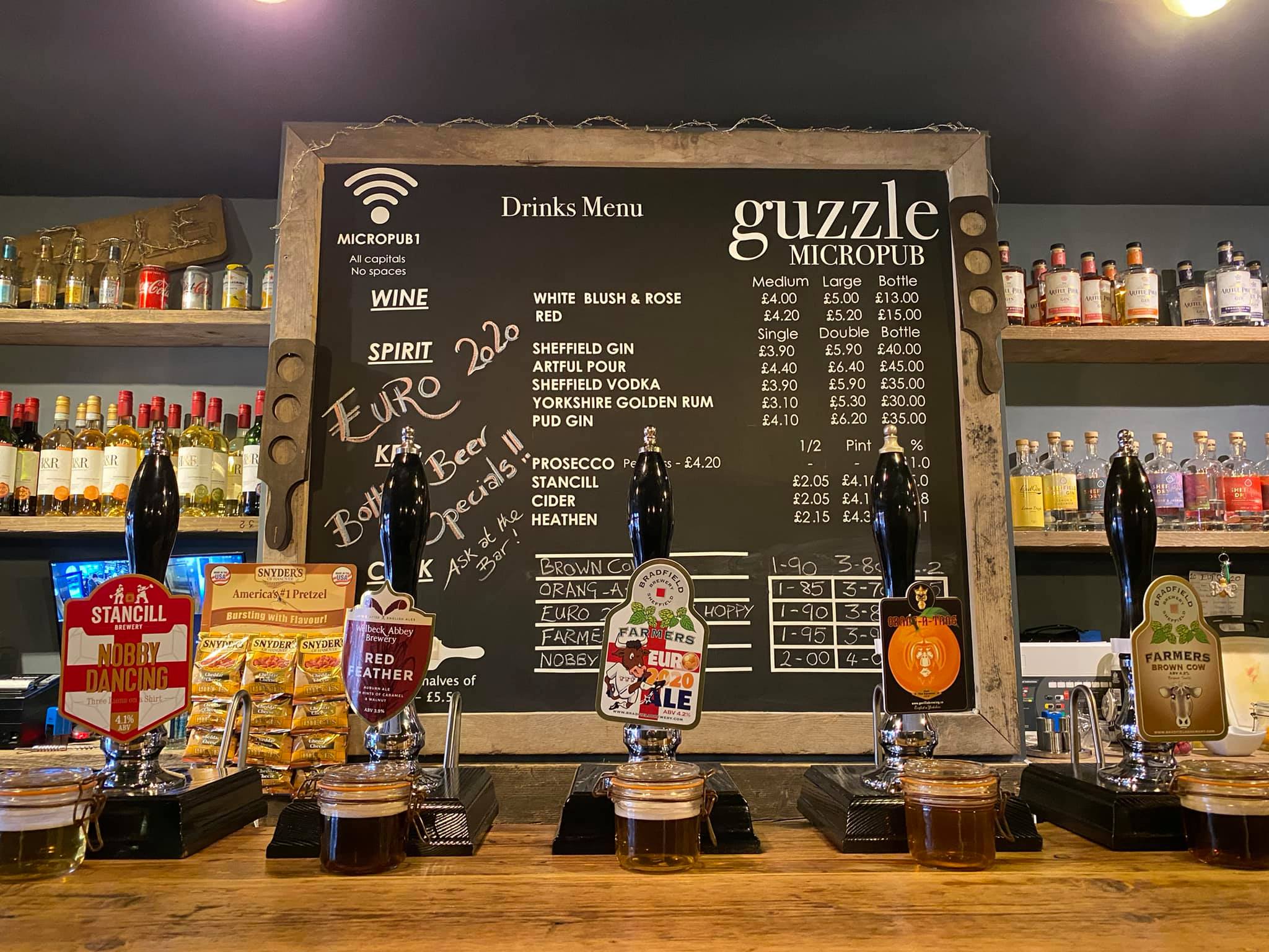 The bar at Guzzle with several beer pumps, a large blackboard with drinks and prices and shelves filled with bottles.
