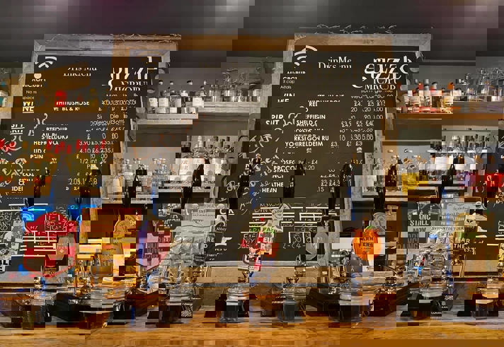 The bar at Guzzle with several beer pumps, a large blackboard with drinks and prices and shelves filled with bottles.