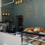 The counter at Kollective Kitchen - Nether Edge with a glass display case full of cakes and pastries. The back wall is a dark sage green with the coffee menu picked out in gold type.