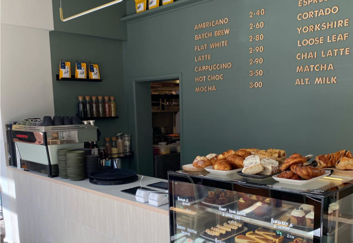 The counter at Kollective Kitchen - Nether Edge with a glass display case full of cakes and pastries. The back wall is a dark sage green with the coffee menu picked out in gold type.