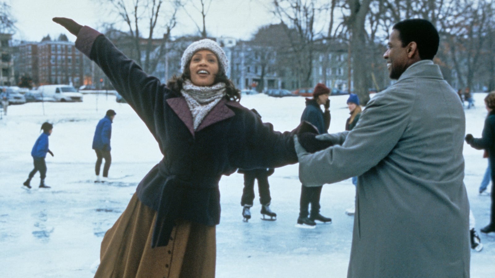 Two people ice skating outdoors on a frozen surface, with one person extending arms gracefully while the other holds their hand for support. Both are dressed in winter clothing, including coats and scarves. In the background, several others are skating and walking on the ice, with snow-covered ground and bare trees surrounding the area. Buildings are visible in the distance under a pale winter sky.
