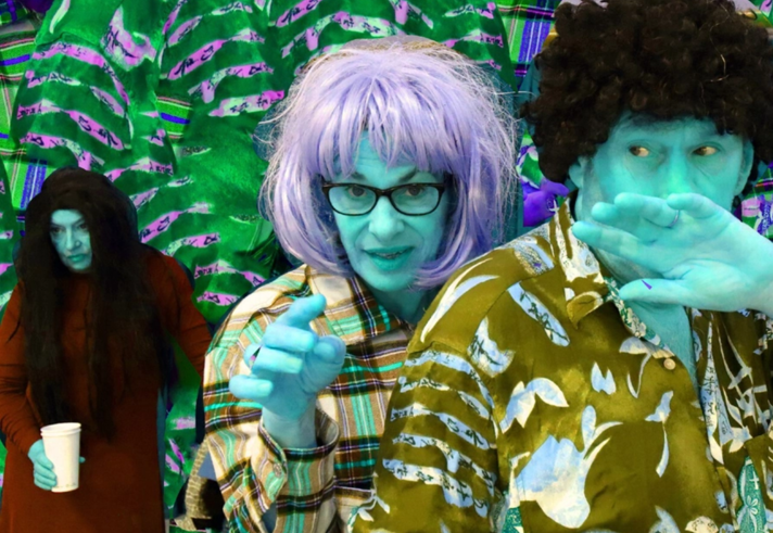 Promotional image for Cold Sweat featuring a man and woman with turquoise skin and contrasting hair colours. They wear colourful patterned shirts. Behind them, a woman with long black hair holds a cup. They stand against a green leafy background.