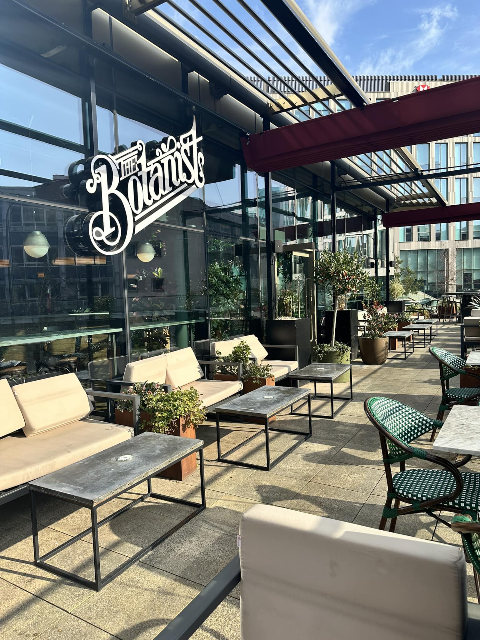 An outdoor terrace with marble-topped tables and green woven chairs, set against a backdrop of modern buildings on a bright, sunny day.
