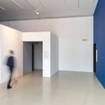 Interior view of SITE Gallery showing a minimalist white hallway with a bright blue feature wall and a person walking past the entrance.