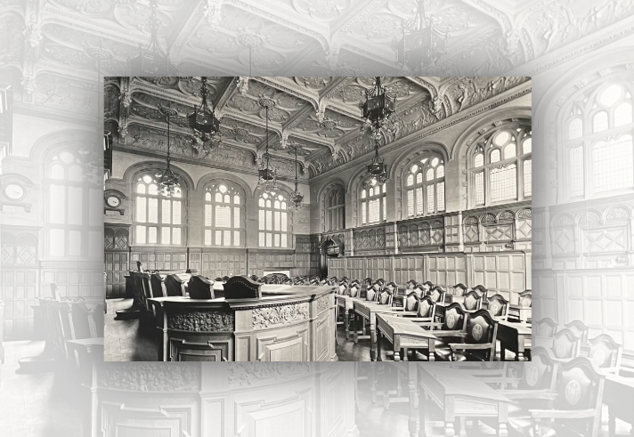 Black-and-white photograph of an ornate council chamber or assembly hall. Rows of carved wooden desks and chairs face a large central table with detailed decorative panels. Tall arched windows line the walls, filling the room with light. The high vaulted ceiling is richly decorated with intricate plasterwork, relief sculptures and hanging lantern-style chandeliers. Wood panelling surrounds the room, and a clock is mounted on the left wall. No people are present.