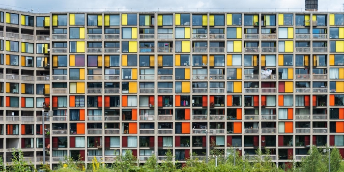 Front view of a large apartment building with a grid-like facade featuring numerous square windows and panels in bright colors, including yellow, orange, and red. The building has multiple balconies and is set against a cloudy sky, with green shrubs and trees in the foreground.