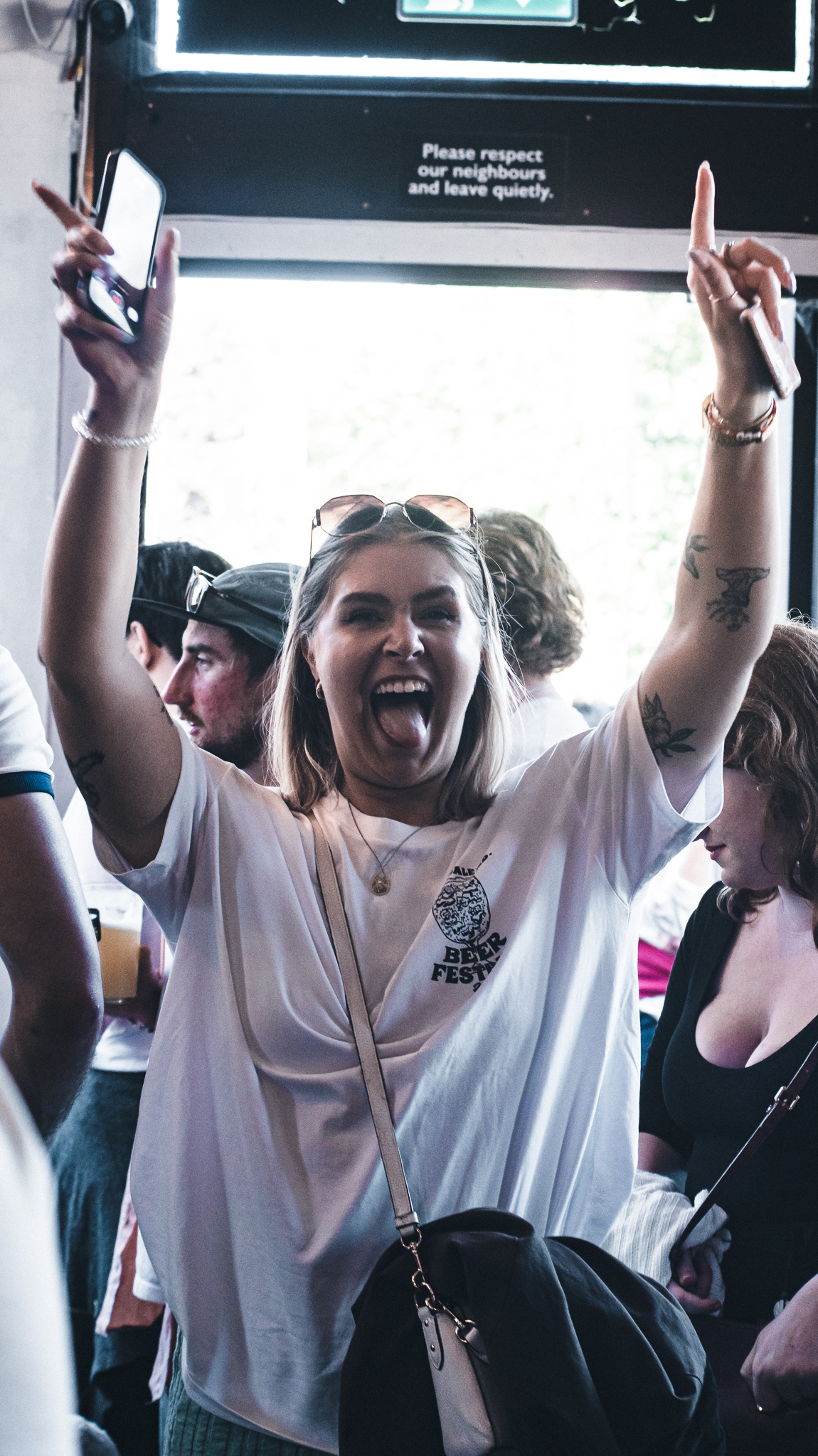 A person in a busy indoor venue raising both arms in the air while holding a phone, with a group of people socialising behind them.