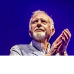 Close-up stage photograph of a person with short white hair and a white beard wearing a light-coloured jacket. The person is shown from the chest up against a vivid blue background, with hands raised together near the face. Bright stage lighting highlights the face and clothing, creating a striking concert or public-event portrait.