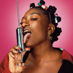 Promotional photograph showing a person against a bright pink background holding a large metal syringe upright beside their face. The person has styled hair with decorative clips and accessories, statement earrings, long patterned nails and a red jacket. The close-up studio portrait uses bold colours and dramatic lighting, creating a striking, contemporary image.