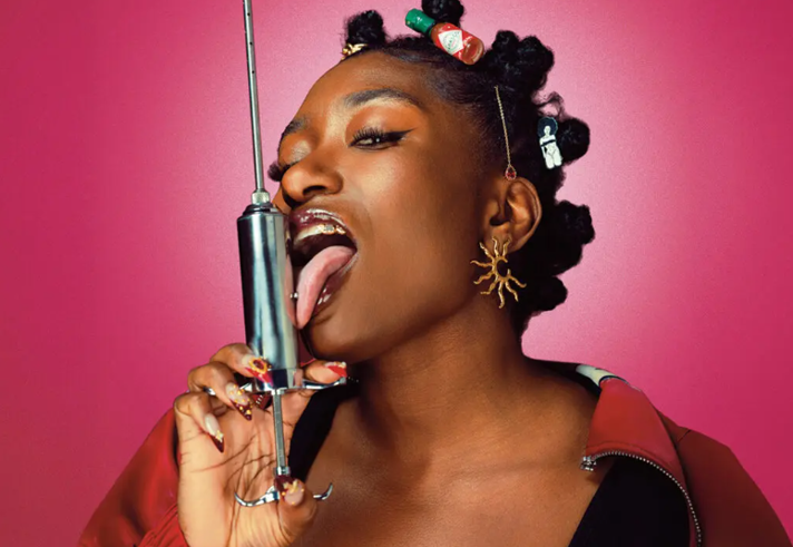 Promotional photograph showing a person against a bright pink background holding a large metal syringe upright beside their face. The person has styled hair with decorative clips and accessories, statement earrings, long patterned nails and a red jacket. The close-up studio portrait uses bold colours and dramatic lighting, creating a striking, contemporary image.