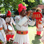 A lively outdoor performance featuring a traditional Indian brass band dressed in ornate white outfits with colourful embroidery and bright red turbans. Musicians play instruments including a trumpet, clarinet, and drums, with a red banner reading “RHBB” on the trumpet. The group stands on grass under trees, with festival stalls and people visible in the background on a sunny day.