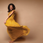 Sindhu Vee in a mustard yellow flowing dress photographed in front of a sepia background.