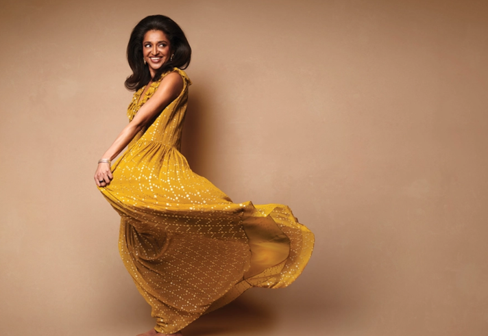 Sindhu Vee in a mustard yellow flowing dress photographed in front of a sepia background.