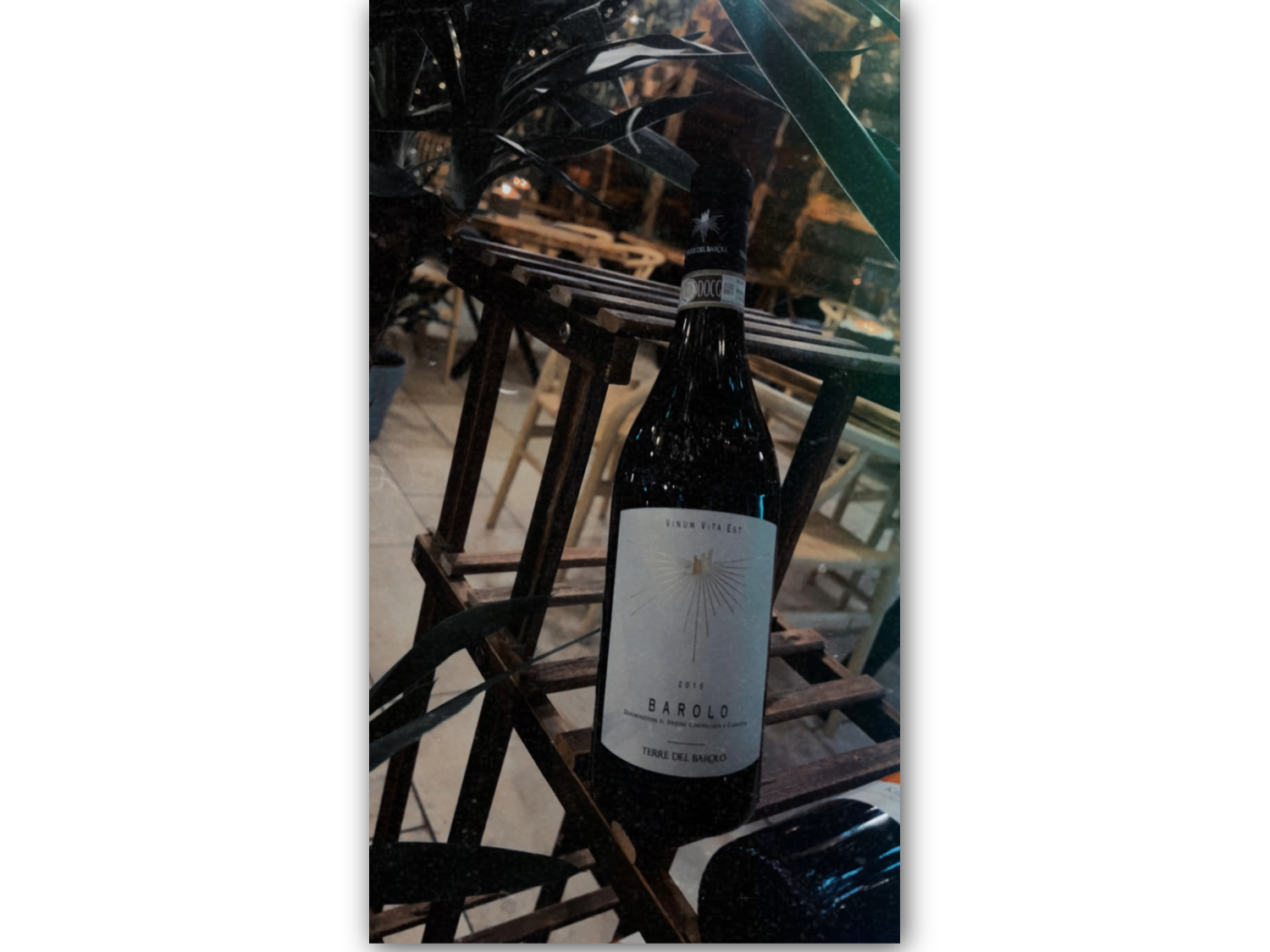 A bottle of 2014 Barolo wine labeled "Vinum Vita Est" and "Terre del Barolo" displayed on a wooden rack. The background features chairs and tables in an indoor dining setting, suggesting a restaurant atmosphere.