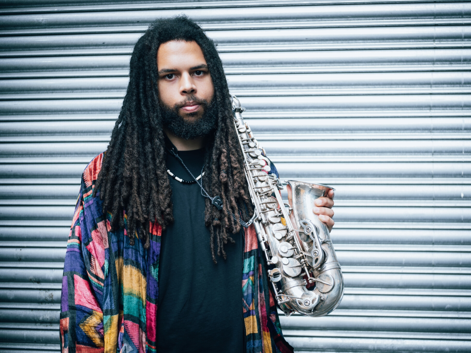 A person with long dreadlocks stands in front of a corrugated metal wall, wearing a colourful patterned jacket over a dark shirt. They hold a silver alto saxophone in one hand, angled slightly forward, with its keys and curved bell clearly visible.