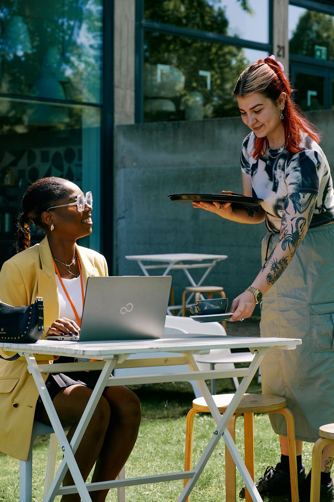 A woman is sat at a table working on a laptop, while another woman brings her a cup of coffee.