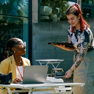A woman is sat at a table working on a laptop, while another woman brings her a cup of coffee.