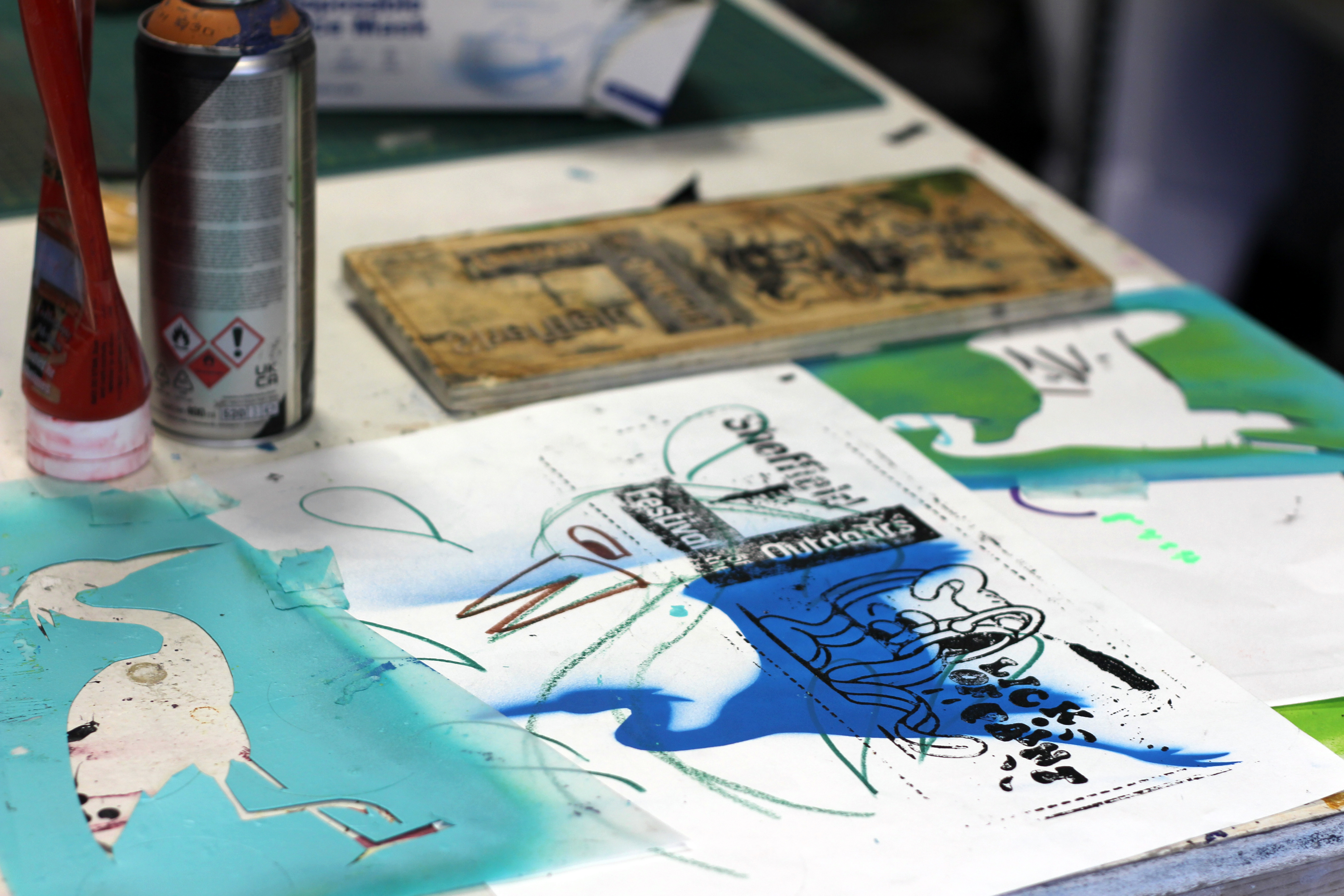 A cluttered art workspace featuring a spray paint can, a red-handled tool, and a wooden block on a table. The surface is covered with colorful abstract designs, including blue and green paint, stylised text, and illustrations of birds.