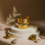 Elegant wide gold cuff bracelet with hammered texture, showcased on stacked stone display with geometric gold accents and dried foliage in soft lighting