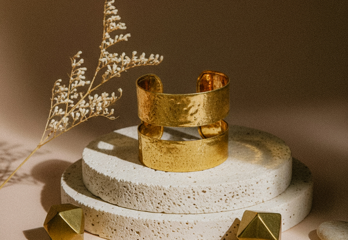 Elegant wide gold cuff bracelet with hammered texture, showcased on stacked stone display with geometric gold accents and dried foliage in soft lighting