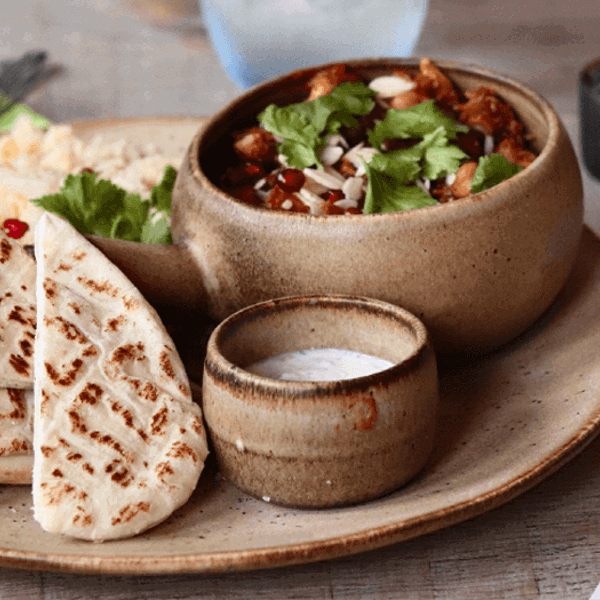 Food at Ego Mediterranean.  A rustic ceramic plate with folded grilled flatbreads, a small bowl of creamy white dip, and a larger bowl of stew topped with fresh herbs and sliced almonds. The plate sits on a wooden table alongside cutlery, a salt and pepper set, and a postcard featuring a coastal scene with turquoise water and a red-and-white boat.